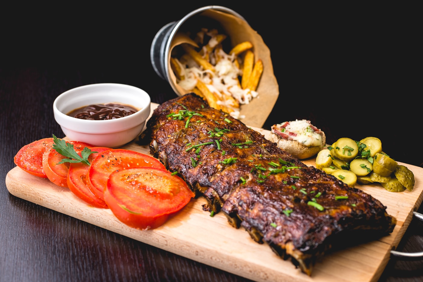 A rack of glazed ribs plated on a wooden board with fries, tomatoes and pickles